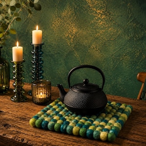 Peut inclure: Nature morte avec une théière en fonte noire sur un dessous de plat en feutre bleu et jaune. Deux bougies allumées dans des bougeoirs en verre décoratifs et un vase avec de la verdure sur une table en bois.