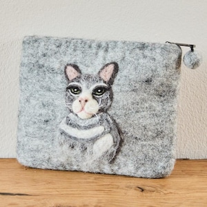 Könnte beinhalten: Eine rechteckige Tasche aus grauem Filz mit einem nadelgefilzten Katzenmotiv auf der Vorderseite. Die Katze hat dunkelgraues und weißes Fell, grüne Augen und eine rosa Nase. Eine kleine graue Filzkugel ist am Metallreißverschluss befestigt.