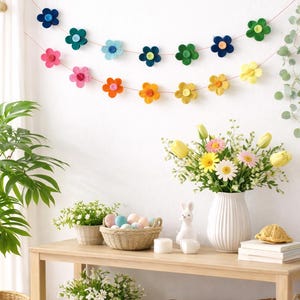 Könnte beinhalten: Eine farbenfrohe Blumengirlande hängt an einer weißen Wand. Darunter zeigt ein Holztisch einen Korb mit Ostereiern, einen weißen Keramikhasen und eine Blumenvase. Die Girlande hat Filzblumen in verschiedenen Farben.