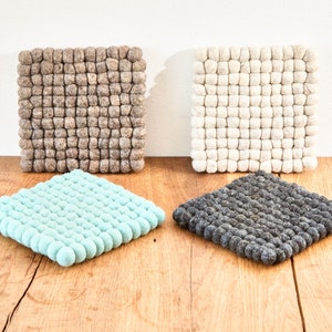Peut inclure: Quatre dessous de verre carrés en feutre dans les tons de gris, blanc et turquoise, disposés sur une surface en bois. Les dessous de verre sont faits de petites boules de feutre arrondies, créant un aspect texturé. Articles de décoration.