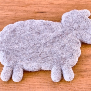 Può includere: Un sottobicchiere di feltro grigio a forma di pecora. La pecora ha un corpo arrotondato e quattro zampe.