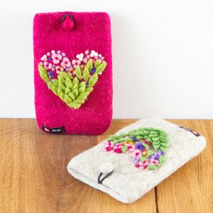 Puede incluir: Dos fundas de teléfono de fieltro con un diseño de corazón floral. La funda de la izquierda es rosa con un corazón verde y morado. La funda de la derecha es blanca con un corazón verde y rosa.