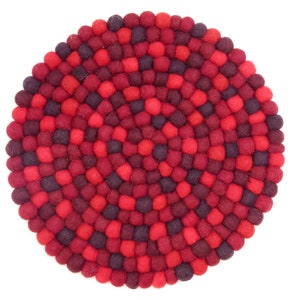 Può includere: Un sottobicchiere rotondo composto da palline di feltro nei toni del rosso e del bordeaux. Le palline di feltro sono disposte a spirale, creando una superficie strutturata. Il sottobicchiere è progettato per proteggere le superfici da calore e umidità.