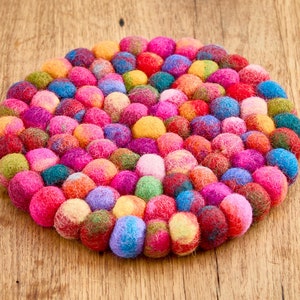 Puede incluir: Un salvamanteles redondo de fieltro compuesto por numerosas bolitas de fieltro. El salvamanteles presenta una vibrante gama de colores, incluyendo rosa, rojo, azul, amarillo y verde. Se encuentra sobre una superficie de madera.