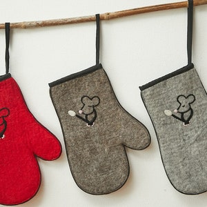 Könnte beinhalten: Drei Ofenhandschuhe hängen an einer Holzstange. Die Handschuhe sind aus Filz und haben einen schwarzen Saum. Jeder Handschuh hat ein kleines gesticktes Mausdesign. Ein Handschuh ist rot, einer ist grau und einer ist braun.