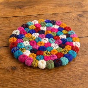Può includere: Un sottopentola rotondo e colorato composto da numerosi piccoli pezzi a forma di zucca infeltrita. Le zucche sono in una varietà di colori, tra cui rosso, arancione, giallo, blu, rosa e bianco. Il sottopentola è posto su una superficie di legno.