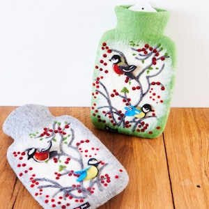 Könnte beinhalten: Zwei Filz-Wärmeflaschen mit winterlichem Design. Beide Flaschen haben einen weißen Hintergrund mit einem grauen oder grünen Filz-Außenmaterial. Das Design beinhaltet einen Ast mit roten Beeren und kleinen Filzvögeln.