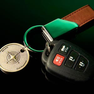 May include: A green and brown leather key chain with a compass charm and a black car key fob with buttons labeled "Hold".