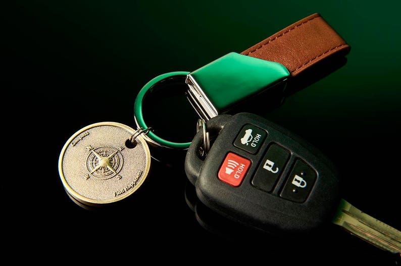 May include: A green and brown leather key chain with a compass charm and a black car key fob with buttons labeled "Hold".