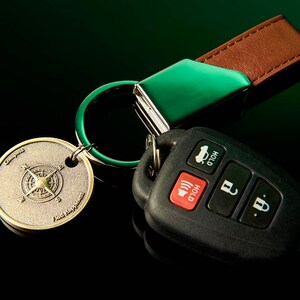 May include: A green and brown leather key chain with a compass charm and a black car key fob with buttons labelled "Hold".