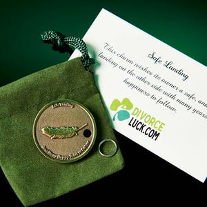 May include: A small, gold-colored charm with a feather design, in a green velvet pouch. The charm is accompanied by a card that reads "Safe Landing" and "DIVORCE LUCK.COM".