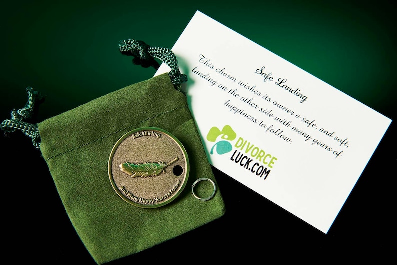 May include: A small, gold-colored charm with a feather design, in a green velvet pouch. The charm is accompanied by a card that reads "Safe Landing" and "DIVORCE LUCK.COM".