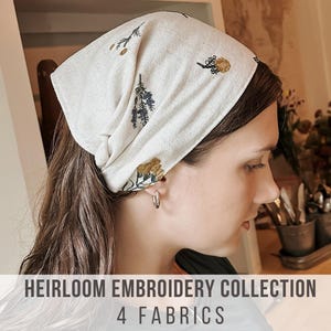 May include: An off-white head wrap featuring a delicate botanical embroidery pattern in shades of gold, green, and dark blue. The fabric is textured and worn tied around the head. The text HEIRLOOM EMBROIDERY COLLECTION 4 FABRICS is visible.