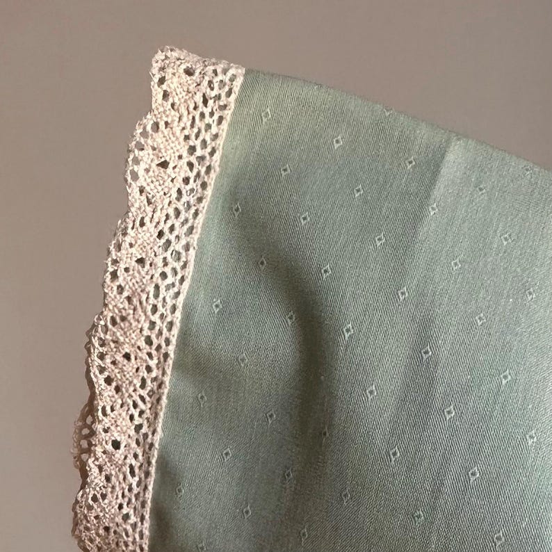 May include: A green fabric with a white lace trim. The fabric has a small, repeating diamond pattern.