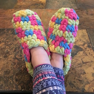 May include: A pair of crocheted slippers in a colorful pattern of pink, blue, yellow, and green. The slippers are worn on a person's feet, with the legs of the person wearing patterned leggings.