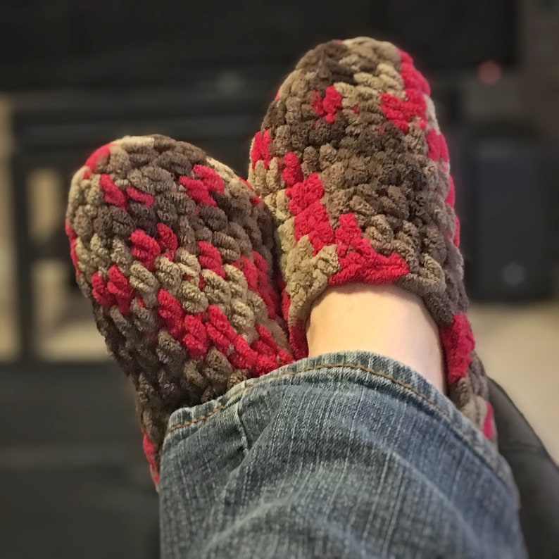 May include: A pair of fuzzy slippers with a camouflage pattern in shades of brown, red, and beige. The slippers are worn on a person's feet, with the toes of the slippers tucked under the person's jeans.