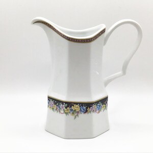 May include: White ceramic pitcher with a hexagonal body and a curved handle. The pitcher features a floral band with blue, yellow, and pink flowers on a black background. A decorative border with a gold and burgundy pattern adorns the rim.