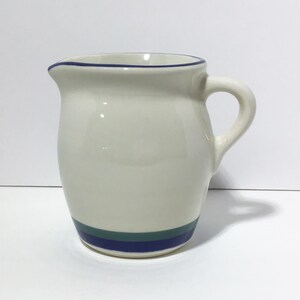 May include: Cream-colored ceramic pitcher with a curved handle and a spout. The pitcher has a blue line around the rim and a blue and teal stripe near the base. The pitcher is a classic design for serving beverages.