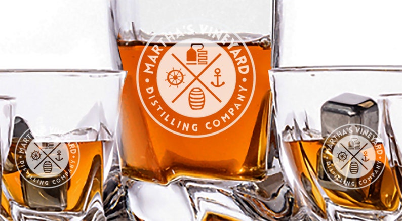 May include: Four clear glass tumblers filled with amber liquid. The tumblers have a logo on the bottom that reads "Martha's Vineyard Distilling Company" with a nautical design.