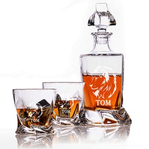 May include: A crystal decanter and two matching glasses with a lion head design and the name "TOM" engraved on each piece. The decanter is filled with amber liquid.