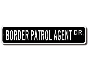 May include: A rectangular black and white street sign that reads "BORDER PATROL AGENT DR." The sign has a white border and is mounted on a white background. The text is in a bold, sans-serif font.