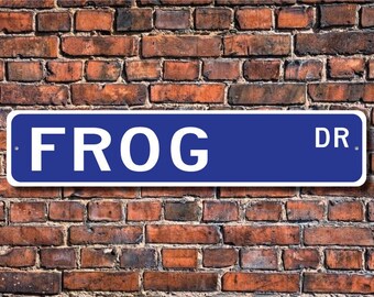 Może przedstawiać: Niebiesko-biały znak drogowy z napisem "FROG DR." na tle ceglanego muru.