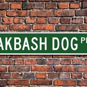 Puede incluir: Un cartel de calle verde y blanco con el texto "AKBASH DOG PL." sobre un fondo de pared de ladrillo.