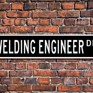 May include: Black and white street sign with the text "Welding Engineer Dr." on a brick wall background.