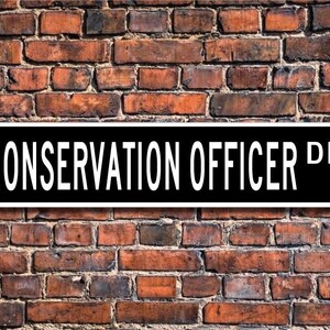 May include: Black and white street sign with the text "CONSERVATION OFFICER DR." against a brick wall background.