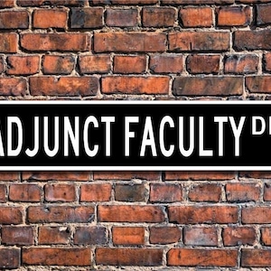 Puede incluir: Un letrero de calle blanco y negro con el texto "ADJUNCT FACULTY DR." sobre un fondo de pared de ladrillo.