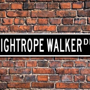Puede incluir: Un letrero de calle blanco y negro con el texto "TIGHTROPE WALKER DR." sobre un fondo de pared de ladrillo.