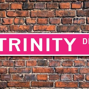Puede incluir: Un cartel de calle rosa con letras blancas que dice "TRINITY DR."
