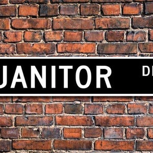 以下が含まれることがあります： 黒と白の街路標識で、黒い背景に白い文字で「JANITOR DR.」と書かれています。