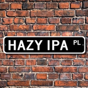 May include: A black and white sign that reads "HAZY IPA PL" is mounted on a weathered brick wall. The sign has rounded corners and the text is in a bold, sans-serif font.