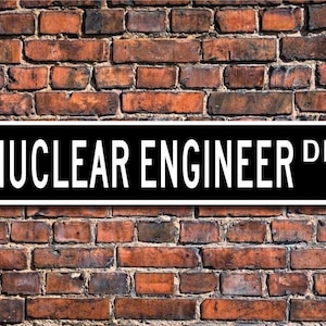 May include: A black and white street sign that reads "NUCLEAR ENGINEER DR" is mounted on a weathered brick wall. The sign is rectangular with rounded corners and the text is in a bold, sans-serif font.