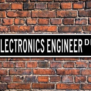 Può includere: Un cartello stradale bianco e nero con il testo "ELECTRONICS ENGINEER DR." su uno sfondo di muro di mattoni.