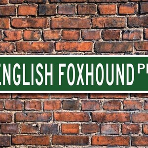 May include: A green street sign with white text that reads "ENGLISH FOXHOUND PL" is mounted on a weathered brick wall. The sign is rectangular with rounded corners. The brick wall is made of red and brown bricks.