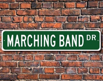 May include: A green street sign with white lettering that reads "MARCHING BAND DR" is mounted on a weathered brick wall. The sign has rounded corners and is centered in the frame. The brick wall is made of red and brown bricks.
