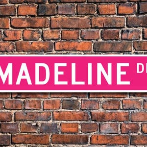 May include: A pink street sign with white lettering that reads "MADEINE DR."