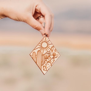 Peut inclure: Une décoration en bois avec un motif découpé au laser représentant une scène de désert avec des cactus, un soleil et des montagnes. La décoration est de couleur brun clair et a une forme de diamant.
