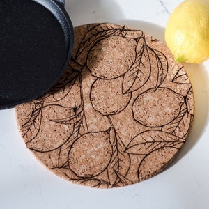 May include: A black cast iron frying pan sits next to a round cork trivet featuring a lemon and leaf pattern. Two yellow lemons are also visible. The items are arranged on a white surface, suggesting a kitchen setting.