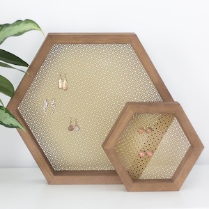 May include: Two hexagonal wooden earring holders with a woven mesh design. The larger holder has several pairs of earrings displayed on it. The smaller holder has one pair of earrings displayed on it. The holders are sitting on a white surface.