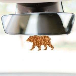 May include: A brown bear-shaped car air freshener hanging from the rearview mirror of a car. The bear is made of a material that looks like wood and has a pattern of words on it.