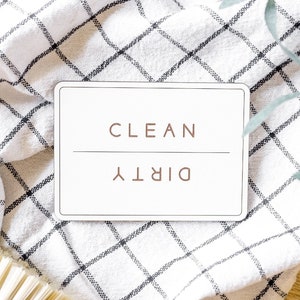 May include: A white rectangular sign with black text that reads "CLEAN" and "DIRTY" on a white and black checkered kitchen towel. A brown brush with beige bristles is in the lower left corner of the image.