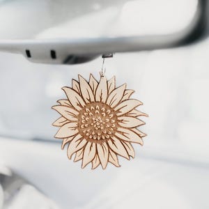 May include: A wooden sunflower car charm hanging from a rearview mirror. The charm is intricately cut with detailed petals and a textured center, showcasing a natural wood grain. The charm adds a touch of nature to the car's interior.
