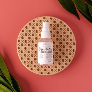 May include: A clear bottle with a white pump dispenser containing rose water and aloe cleanser. The bottle is on a round, woven, light brown coaster. The background is a solid pink.
