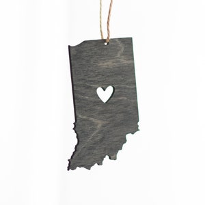 May include: A wooden ornament shaped like the state of Indiana with a heart cut out in the center. The ornament is painted gray and has a rustic finish.