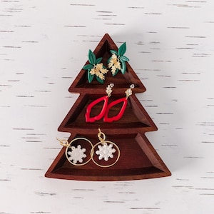May include: A wooden Christmas tree-shaped display stand with three tiers, featuring a set of red, white, and gold earrings. The earrings are arranged in a decorative pattern on the stand.