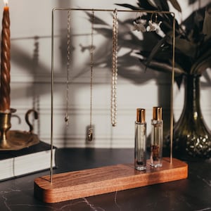 May include: A wooden jewelry stand with a gold metal frame. The stand has three necklaces hanging from it, two gold and one pearl. There are two small bottles on the stand, one clear and one with a brown liquid.