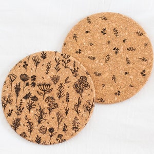 May include: Two round cork trivets with a black floral pattern. The trivets are on a white background.